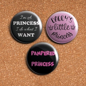 May include: Three round black and pink buttons with white and pink text. The buttons read "I'm a Princess I do what I WANT", "Daddy's little princess" and "Pampered Princess".