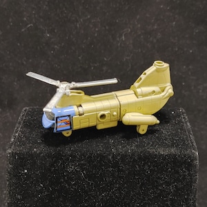 Könnte beinhalten: Ein Spielzeughelikopter in beige und silberner Farbgebung. Der Helikopter hat einen blauen und orangenen Streifen an der Seite. Der Helikopter steht auf einer schwarzen Oberfläche.