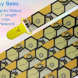 Op de afbeelding: Een geel en zwart grosgrain lint met een bijen- en honingraatpatroon. Het lint is 10-12 inch lang en heeft een gele KAM-clip. Het lint is gelabeld met "Busy Bees".