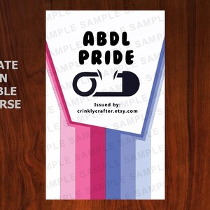 K&ouml;nnte beinhalten: Ein wei&szlig;es rechteckiges Design mit den schwarzen Worten "ABDL PRIDE" und einer Grafik einer Windel-Sicherheitsnadel. Das Design hat rosa und blaue Streifen und den Text "ALTERNATE DESIGN AVAILABLE ON REVERSE". Der Etsy-Shop ist crinklycrafter.etsy.com.