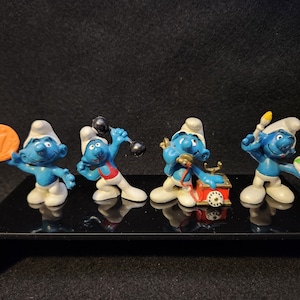 Puede incluir: Cuatro figuritas azules de Pitufo con sombreros blancos. Cada Pitufo sostiene un objeto diferente: una moneda de oro, una mancuerna, un teléfono rojo y una paleta de pintura.