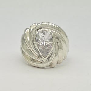 May include: A silver ring with a pear-shaped, clear gemstone at the center. The ring's band features a swirling, textured design around the gemstone. The ring is set against a plain white background.