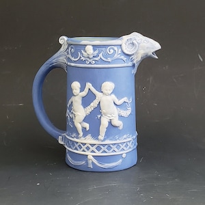 May include: A blue and white ceramic pitcher with a spout shaped like a ram's head. The pitcher features a relief design of two cherubs dancing.