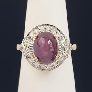 Puede incluir: Un anillo de plata con una gran gema ovalada de color rojo intenso, rodeada por un halo de pequeños diamantes brillantes. El anillo tiene detalles intrincados y pequeños acentos dorados.