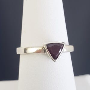 May include: A silver ring with a triangular, dark red gemstone. The gemstone is set in a bezel setting, and the band is smooth and polished. The ring is displayed on a white surface against a dark background.