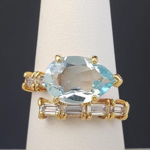 Op de afbeelding: Een gouden ringenset met een peervormige, lichtblauwe edelsteen en een band met rechthoekige, heldere edelstenen. De ring wordt gepresenteerd op een witte standaard tegen een donkere achtergrond.