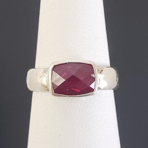 May include: A silver ring with a large, rectangular, faceted, deep red gemstone. The ring is set in a simple bezel setting, and the band has a textured, hammered appearance. The gemstone is the focal point, catching the light and providing a bold pop of color.