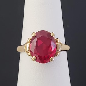 Pode incluir: Um anel de ouro com uma grande pedra preciosa vermelha escura de corte oval. A pedra preciosa é engastada numa banda de ouro com detalhes decorativos. O anel é exibido num suporte branco sobre um fundo preto.