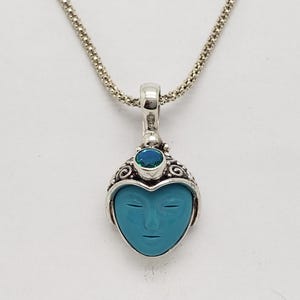 May include: A silver necklace with a turquoise heart-shaped pendant. The pendant has a carved face and a small blue gemstone. The necklace hangs against a white background. The chain is delicate.