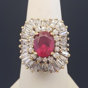 May include: A gold-toned ring featuring a large, oval-cut ruby center stone. The ruby is surrounded by a halo of smaller, round diamonds and baguette-cut stones, creating a radiant, eye-catching design. The ring is set on a white stand against a dark background.