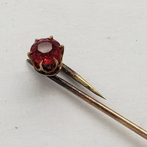 May include: A gold pin with a red gemstone set in a prong setting.