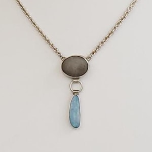 Könnte beinhalten: Eine silberne Halskette mit Kette und Anhänger. Der Anhänger hat einen grauen ovalen Stein über einem hellblauen, tropfenförmigen Stein. Die Halskette wird auf einer weißen Schaufensterpuppe präsentiert.