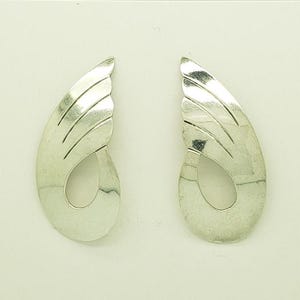 May include: A pair of silver-toned earrings. Each earring has a curved, wing-like shape with etched lines and a teardrop-shaped cutout. The earrings are displayed on a white surface.