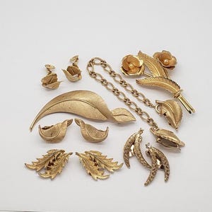 Puede incluir: Una colección de joyas vintage en tono dorado, que incluye broches, pendientes y un collar. Las piezas presentan diseños florales y de hojas, con una variedad de texturas y formas. Las joyas están dispuestas sobre una superficie blanca.