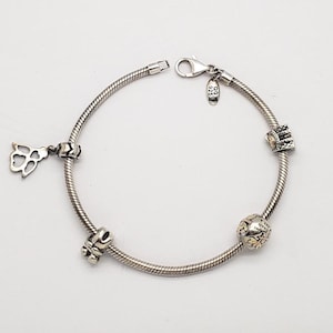 Könnte beinhalten: Ein silbernes Armband mit vier Charms, darunter ein Herz, eine Blume, eine Kugel und eine Krone. Das Armband ist aus einer Schlangenkettenform gefertigt.