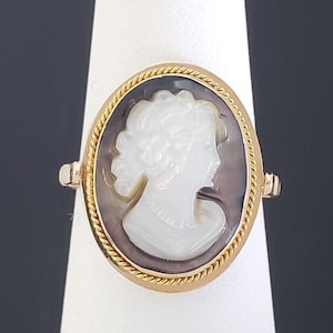 May include: Gold-tone ring featuring an oval cameo with a white profile against a dark background. The ring has a twisted rope-like border and is set on a white display stand. The ring's design is classic and elegant.