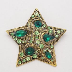 May include: A gold-toned star-shaped brooch with intricate filigree detailing. The brooch is adorned with various sizes of green gemstones, including emerald-cut and teardrop-shaped stones, creating a sparkling effect. The star has five points and a pin closure on the back.