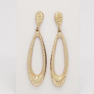 May include: A pair of gold-colored, textured teardrop earrings. The earrings have a textured oval shape with a small, textured top. The earrings are displayed on a white background.