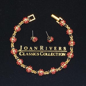 May include: A gold-toned bracelet and matching earrings set. The bracelet and earrings feature red, spherical accents with gold detailing and black beads. The set is presented on a black surface, with the text "JOAN RIVERS CLASSICS COLLECTION" visible.