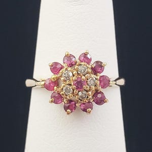 May include: A gold ring featuring a floral design. The ring's center is adorned with small, clear stones, surrounded by a ring of vibrant pink gemstones. The ring is displayed on a white stand against a dark background.