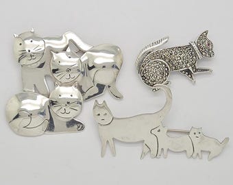 May include: Three silver-toned cat-themed brooches. One brooch features a cluster of four stylised cat faces with smooth, polished surfaces. Another is a single cat with a textured, sparkling finish. The third depicts a cat family of three.