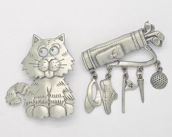May include: Two silver-toned metal brooches. One brooch is a cartoon-style cat with wide eyes and textured fur. The second brooch is a golf bag with dangling charms including a visor, a golf shoe, a flag with the number 4, a tee, and a golf ball.