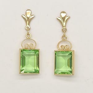 May include: A pair of gold drop earrings featuring rectangular, emerald-cut light green gemstones. Each earring has a decorative gold filigree loop above the stone and a small, stylised gold fleur-de-lis topper attached to a thin gold link.