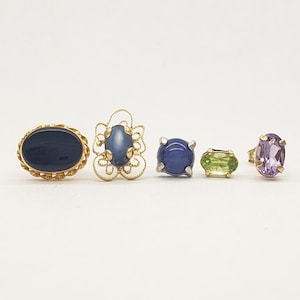 Op de afbeelding: Vijf gouden ringen met verschillende gekleurde edelstenen. De ringen hebben een blauwe ovale steen, een blauwe ronde steen, een groene ovale steen, een paarse ovale steen en een blauwe ovale steen met een decoratieve gouden filigraan setting.