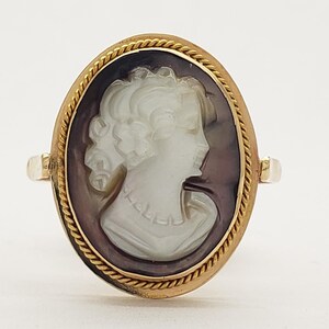 May include: Oval gold ring featuring a cameo of a woman's profile. The cameo is white against a dark background, set within a gold frame with a rope-like border. The ring has a classic, vintage aesthetic.