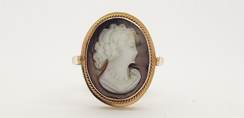 May include: Oval gold ring featuring a cameo of a woman's profile. The cameo is white against a dark background, set within a gold frame with a rope-like border. The ring has a classic, vintage aesthetic.