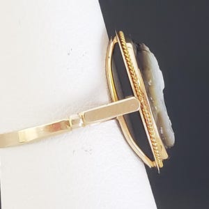May include: A gold-toned ring featuring a cameo with a profile carving. The cameo is set against a black background and framed with a gold border. The ring's band is a simple, polished design.