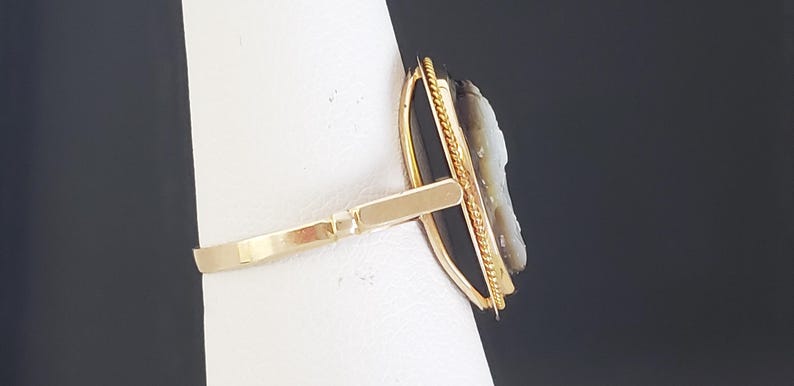 May include: A gold-toned ring featuring a cameo with a profile carving. The cameo is set against a black background and framed with a gold border. The ring's band is a simple, polished design.