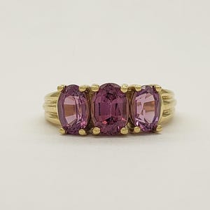 May include: A gold ring featuring three oval-cut, pink gemstones. The gemstones are set in a row, with a ribbed gold band. The ring is displayed on a white background, showcasing the jewelry's design.