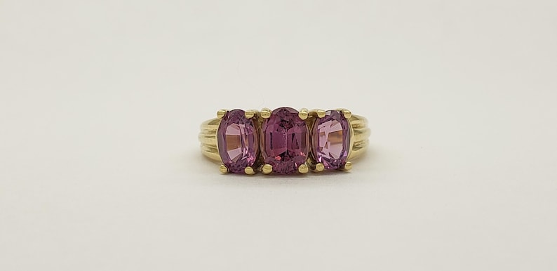 May include: A gold ring featuring three oval-cut, pink gemstones. The gemstones are set in a row, with a ribbed gold band. The ring is displayed on a white background, showcasing the jewelry's design.
