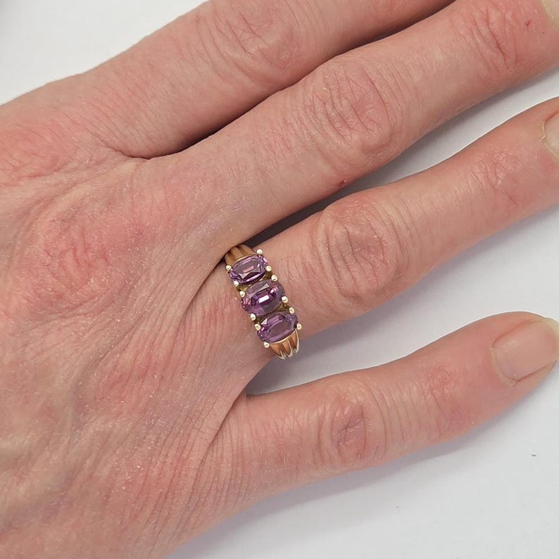 May include: A gold ring featuring three oval-cut purple gemstones. The ring's band is a warm gold color, and the gemstones are set in a row. The ring is shown on a finger against a white background.