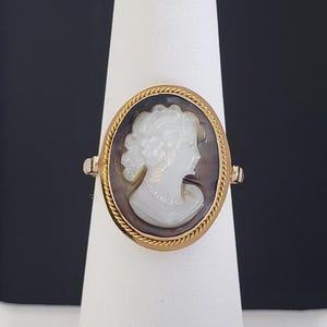 May include: A gold ring featuring an oval cameo. The cameo depicts a white profile of a woman against a dark background, framed by a gold rope-like border. The ring is displayed on a white stand against a dark background.