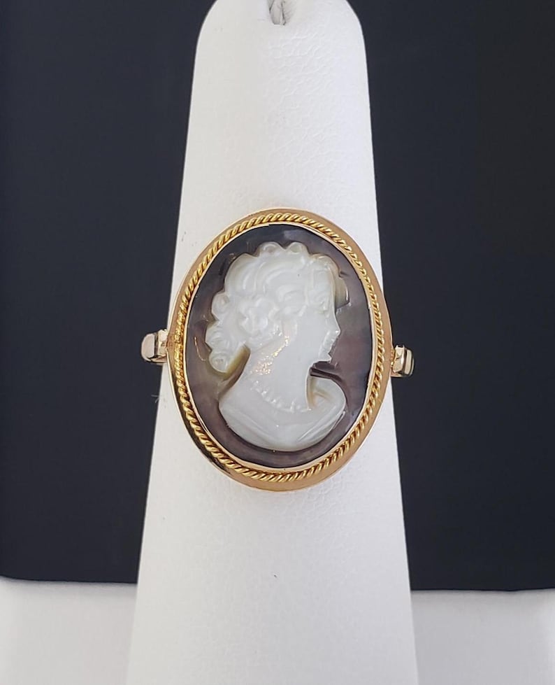 May include: A gold ring featuring an oval cameo. The cameo depicts a white profile of a woman against a dark background, framed by a gold rope-like border. The ring is displayed on a white stand against a dark background.
