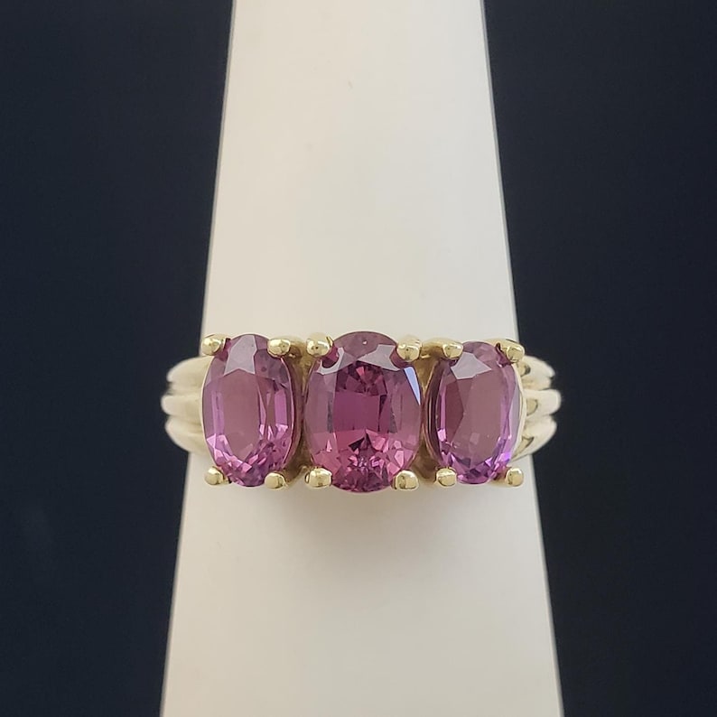 May include: A gold ring featuring three oval-cut, vibrant pink gemstones. The gemstones are set in a row, with a gold band that has a ribbed design on the sides. The ring is displayed on a white stand against a dark background.