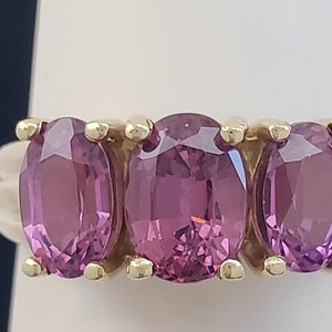 May include: A gold ring featuring three oval-cut, pink gemstones. The gemstones are set in a row, with a gold band and accents. The ring is displayed on a white surface against a dark blue background.