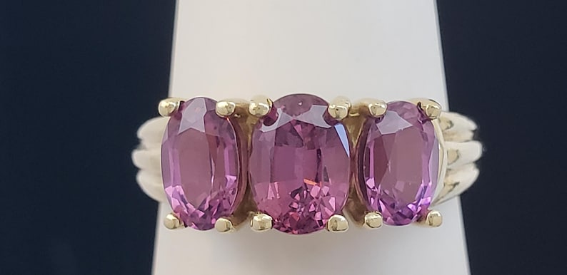 May include: A gold ring featuring three oval-cut, pink gemstones. The gemstones are set in a row, with a gold band and accents. The ring is displayed on a white surface against a dark blue background.