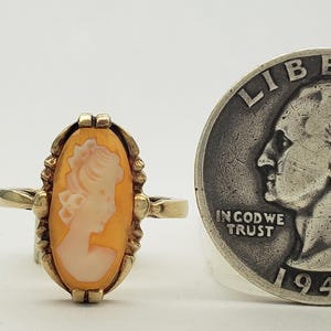 May include: A gold ring with a cameo featuring a woman's profile in a white cameo set in an orange shell. The ring is next to a 1943 US quarter for size comparison.