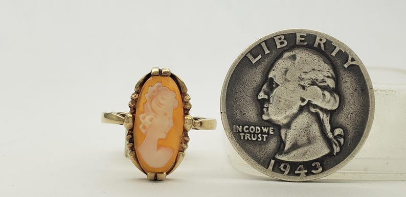 May include: A gold ring with a cameo featuring a woman's profile in a white cameo set in an orange shell. The ring is next to a 1943 US quarter for size comparison.