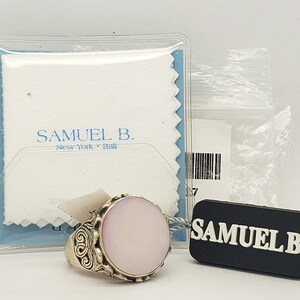 May include: A silver ring with a large oval-shaped pink stone set in a decorative silver setting. The ring is displayed on a white cloth with the text "Samuel B. New York - Italia" printed on it. The ring is accompanied by a black tag with the text "Samuel B."