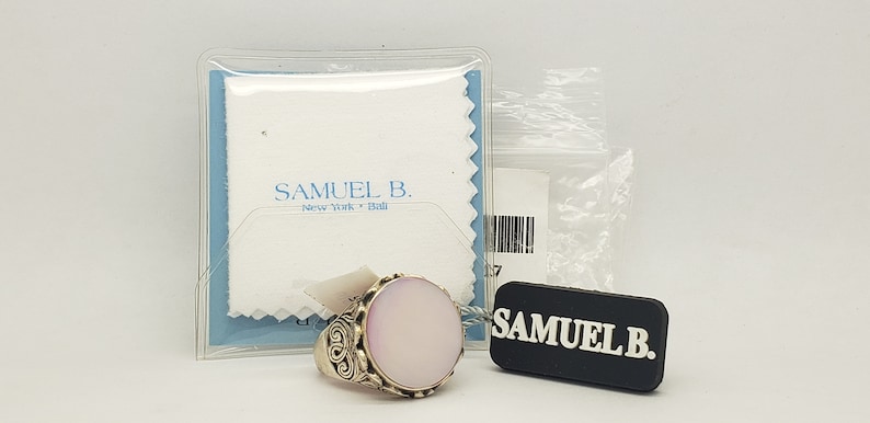 May include: A silver ring with a large oval-shaped pink stone set in a decorative silver setting. The ring is displayed on a white cloth with the text "Samuel B. New York - Italia" printed on it. The ring is accompanied by a black tag with the text "Samuel B."