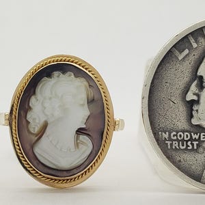May include: An oval gold ring with a cameo of a woman's profile in white against a dark background. A silver quarter coin is next to the ring.