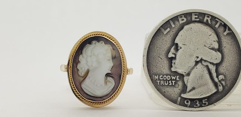 May include: An oval gold ring with a cameo of a woman's profile in white against a dark background. A silver quarter coin is next to the ring.