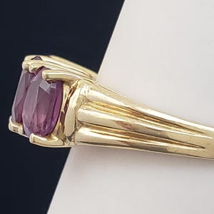 May include: A gold ring featuring two oval-cut, deep purple gemstones. The ring's band is a polished gold, with a ribbed design on the sides. The gemstones are set close together, creating a bold and elegant look.