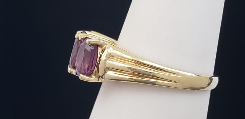May include: A gold ring featuring two oval-cut, deep purple gemstones. The ring's band is a polished gold, with a ribbed design on the sides. The gemstones are set close together, creating a bold and elegant look.