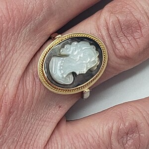 May include: Oval gold-tone ring featuring a cameo design. The cameo depicts a woman's profile in white against a dark background, framed by a twisted rope-like border. The ring is worn on a finger.