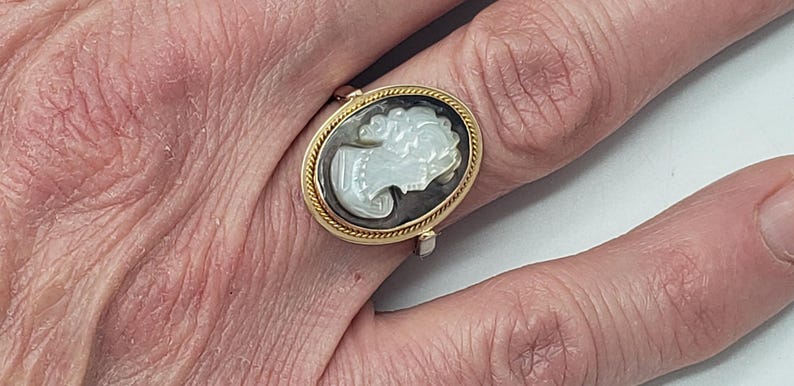 May include: Oval gold-tone ring featuring a cameo design. The cameo depicts a woman's profile in white against a dark background, framed by a twisted rope-like border. The ring is worn on a finger.